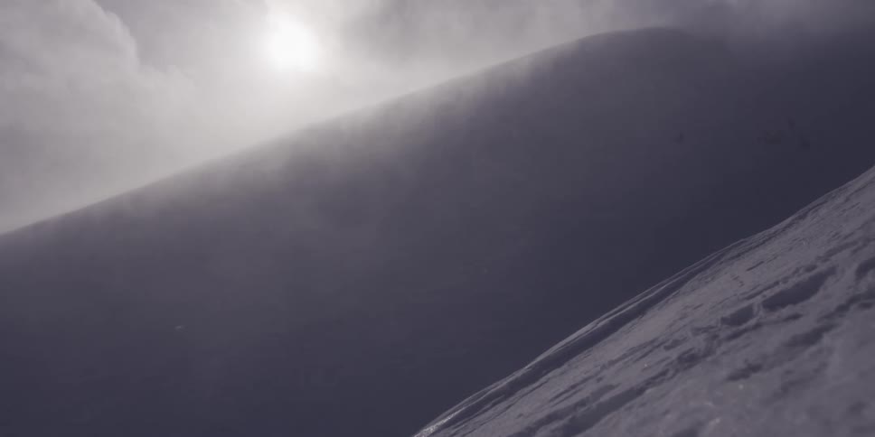 A dramatic slow motion shot of snow blowing across a mountain ridge with the sun shining through the clouds