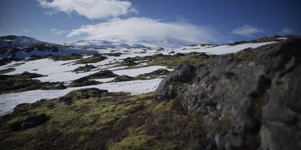 Vast, snowy Icelandic tundra with mossy volcanic rocks stretches out under a bright blue sky with wispy clouds