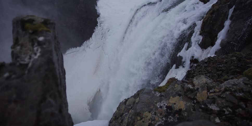 A slow-motion dolly shot reveals a powerful waterfall crashing down over snow-covered rocks