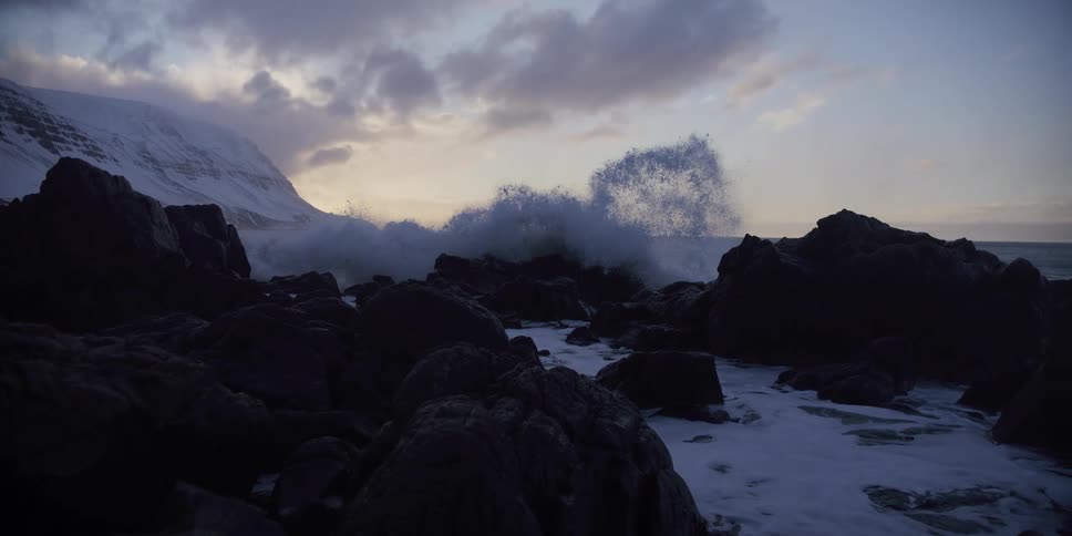 A dramatic slow-motion shot captures a powerful ocean wave exploding against a dark, rocky shore next to a snow-covered mountain at dusk