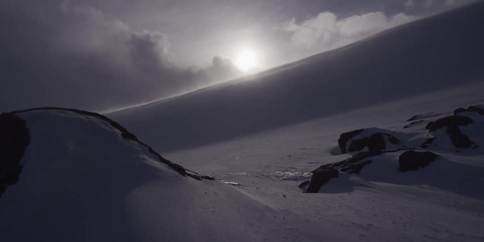 Ethereal sunlight shines over a vast, windy, and snow-covered mountain landscape