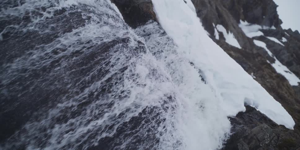 Slow-motion close-up of a powerful waterfall cascading over dark rocks beside a melting snowbank