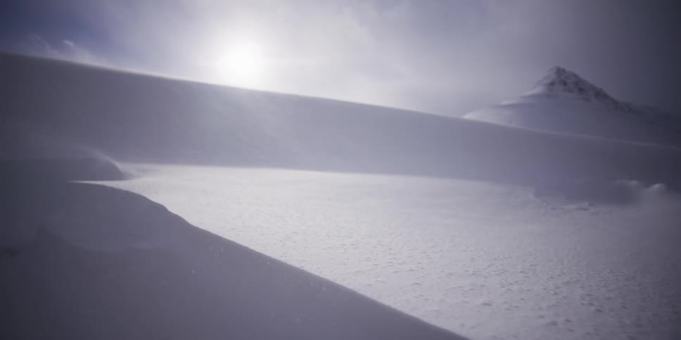 Snow blows across a vast, desolate, and beautiful mountain landscape as the sun shines through the clouds