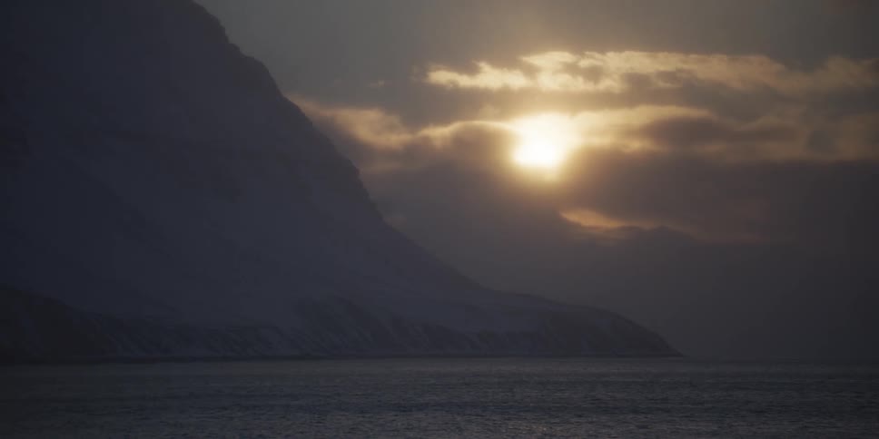 The sun glows behind moody clouds over a vast, snow-covered mountain landscape next to a tranquil sea