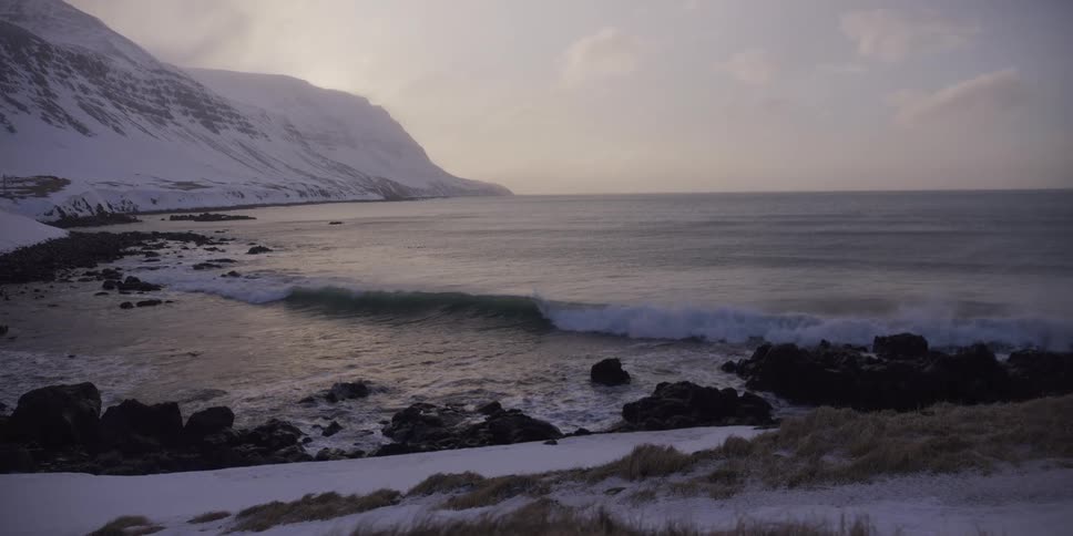 Slow motion footage captures a powerful ocean wave breaking on a frigid, rocky shore in front of snow-covered mountains