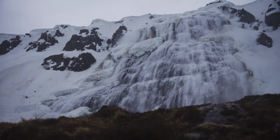 A powerful, tiered waterfall cascades down a snowy and icy mountain slope on a cold, overcast day