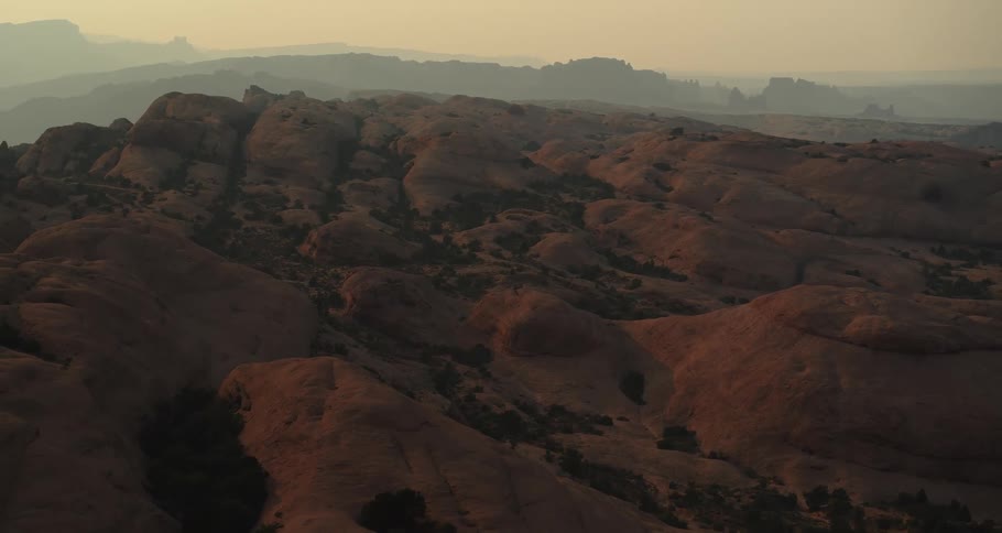 A static wide view of a hazy, golden sunset over the undulating red rock formations of a desert landscape