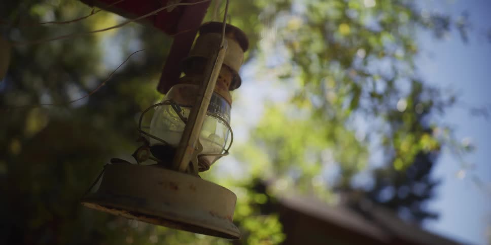 A weathered antique oil lamp hangs from a beam in a rustic outdoor setting with sun-dappled trees in the background