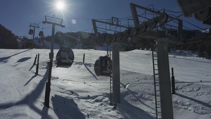 An Aspen ski gondola lift operates on a bright sunny day, with lift cars passing by and skiers descending the snowy mountain in the background