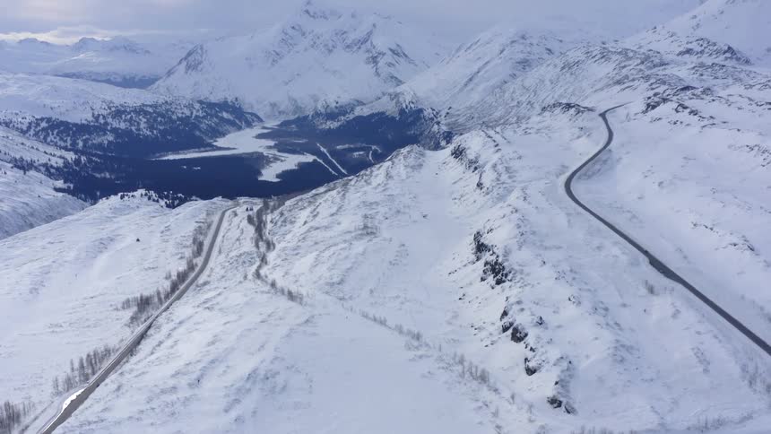 A dramatic aerial view pulls back to reveal a vast, snow-covered mountain pass with winding roads on an overcast winter day