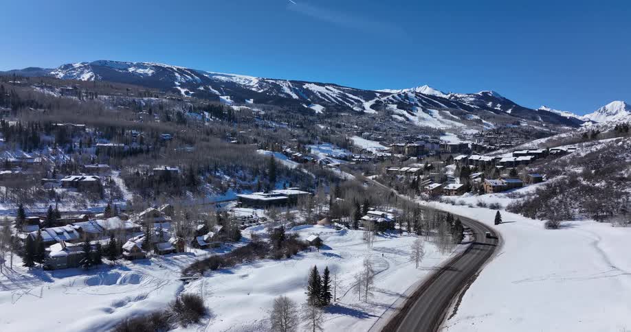 An ascending aerial shot captures a snowy mountain village with a winding road and a ski resort in the background on a sunny day