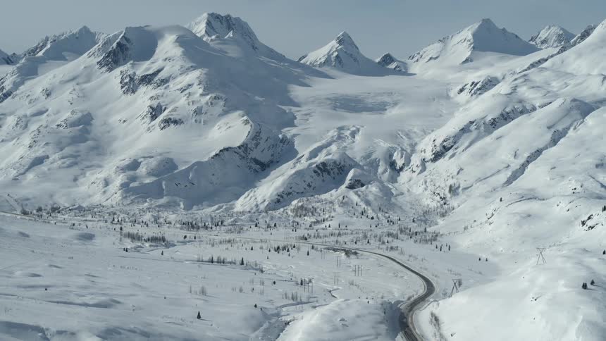 Sweeping aerial view of a remote Alaskan mountain valley covered in snow with a winding road leading towards a glacier