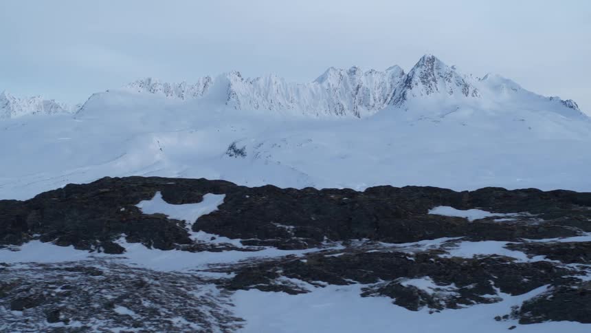 A slow aerial tracking shot passes over a rocky, snow-covered landscape with a massive mountain range in the distance