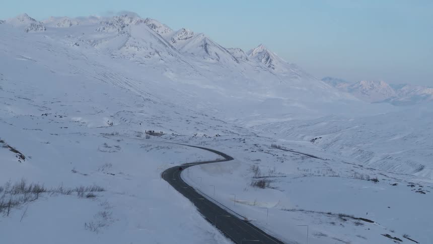 Aerial view of a lone car driving on a scenic, winding road through a vast, snow-covered mountain landscape