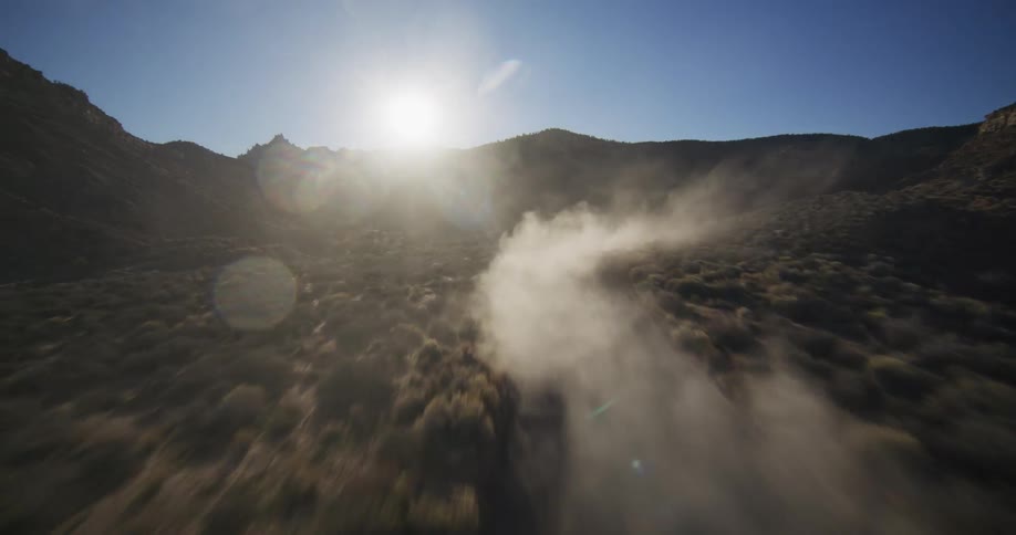 An FPV drone chases an off-road vehicle as it kicks up a massive dust cloud while racing through a desert canyon into the sun