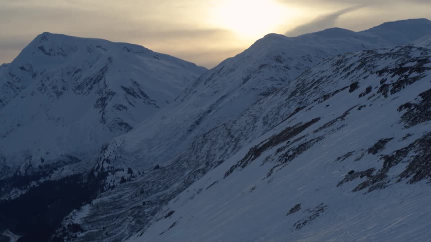A slow aerial pull-back reveals a majestic snow-covered mountain range basking in the soft glow of a winter sunset