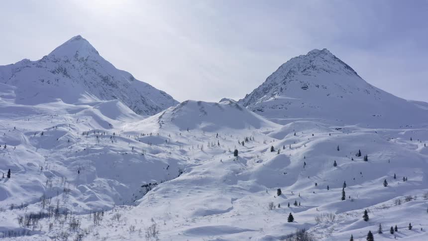 An expansive aerial shot slowly pulls back over a breathtaking, snow-covered mountain wilderness on a sunny day