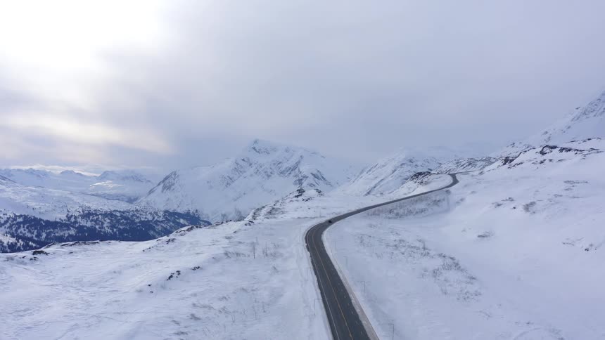 An aerial view of a car driving on a winding road through a vast, snow-covered mountain landscape in winter