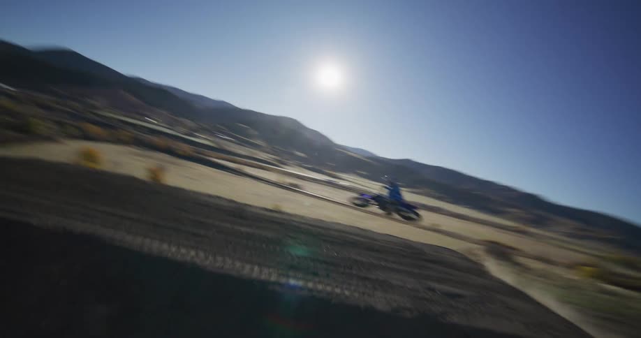 A dynamic aerial shot follows a motocross rider in slow motion as they launch off a large dirt jump in a hilly, rural landscape