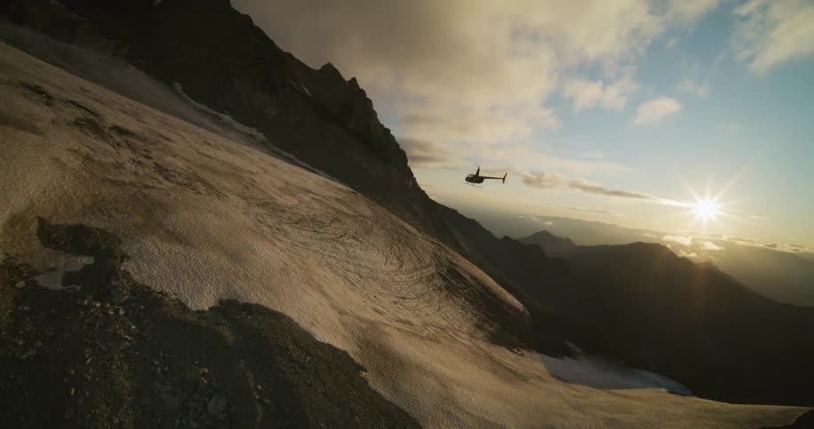 A cinematic slow-motion aerial shot shows a helicopter flying alongside a vast, snow-covered mountain at sunset