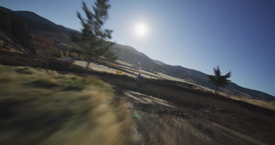 A dynamic FPV drone chases a motocross rider down a dusty dirt track through a beautiful mountain valley on a sunny day