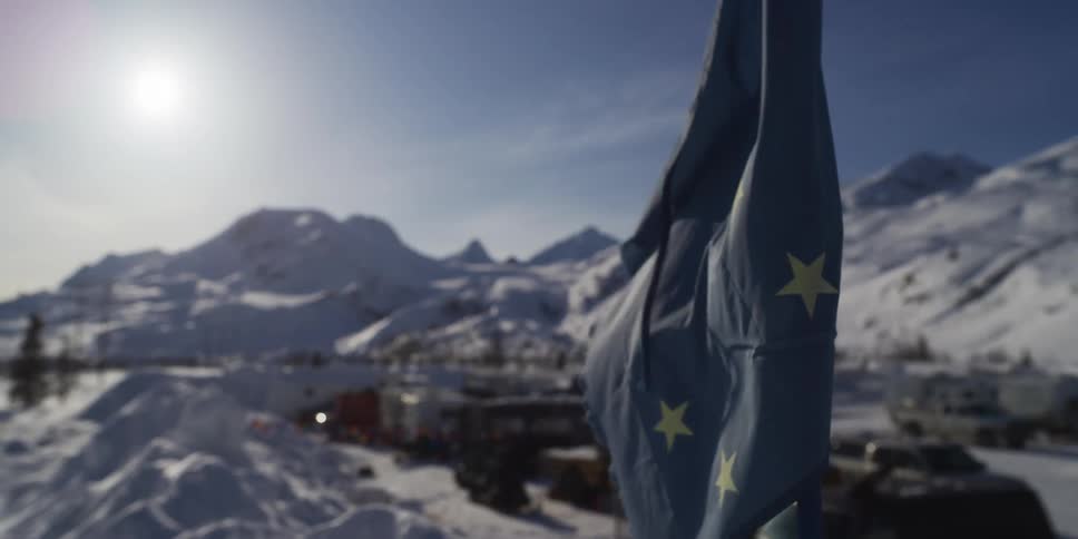 The Alaskan state flag flutters in the bright sun with a beautiful, snowy mountain town in the background