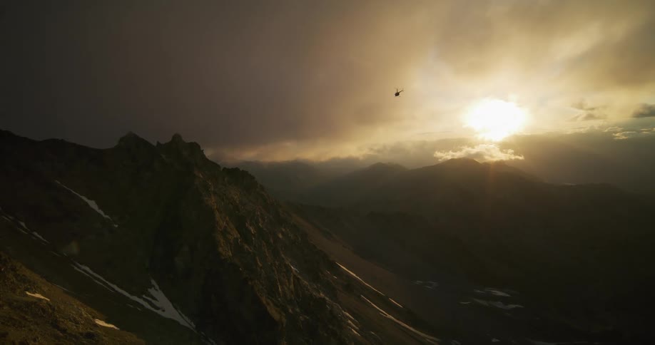 A solitary helicopter flies over a dramatic, rugged mountain range as the golden sun sets behind moody clouds