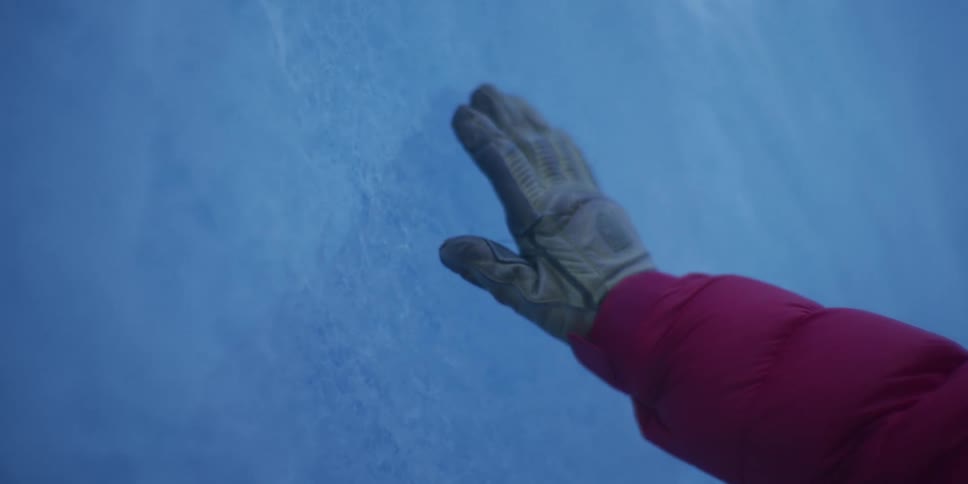 A person's gloved hand gently touches the vast, textured wall of a blue ice glacier