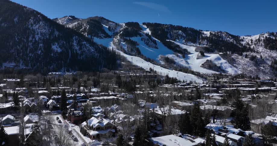 Aerial pullback shot revealing a snow-covered ski resort town nestled at the base of a mountain on a sunny winter day