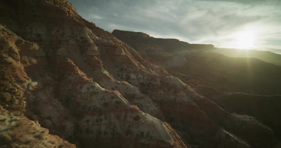 An aerial shot pulls back from a rugged, sunlit mountainside to reveal a vast desert canyon landscape at sunrise
