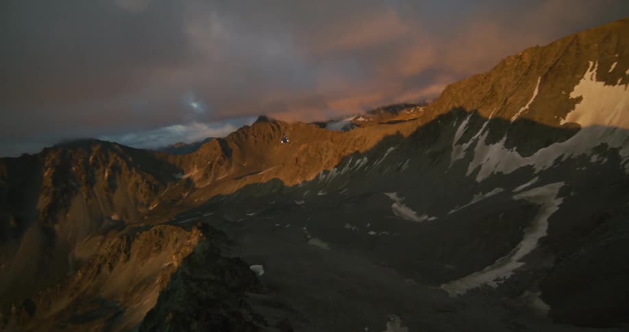 A majestic aerial view shows a helicopter flying past golden-lit peaks in a dramatic mountain range at dusk