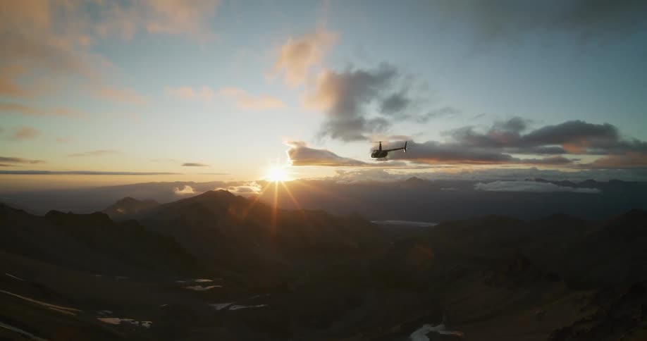 An epic aerial shot follows a helicopter as it flies over a vast mountain range into the golden sunset