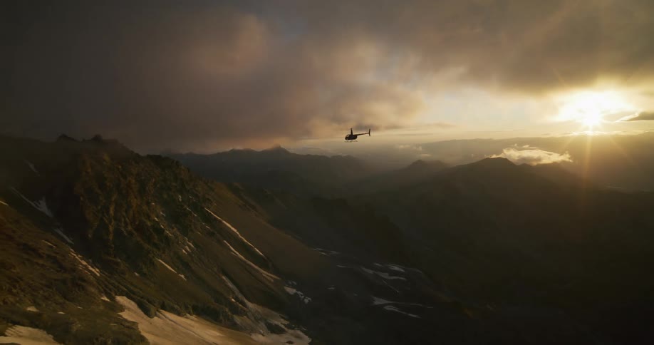 A silhouetted helicopter soars over a majestic, snow-capped mountain range during a breathtaking golden hour sunset