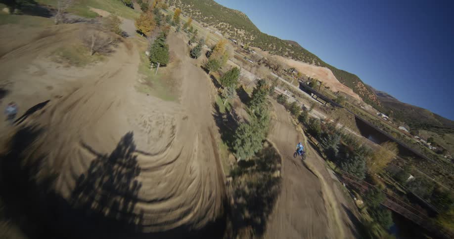 Aerial first-person view of a motocross rider racing down a winding dirt track in the mountains on a sunny day
