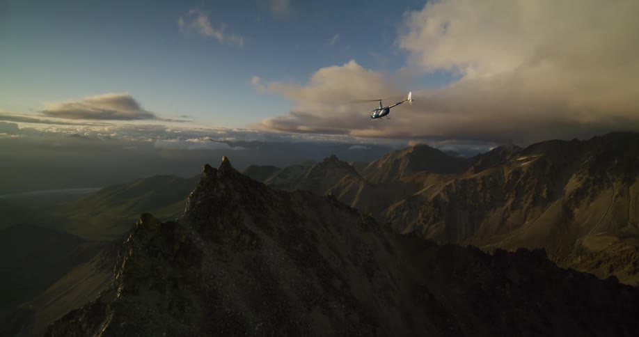 A helicopter flies majestically over a dramatic mountain range during a golden hour sunset