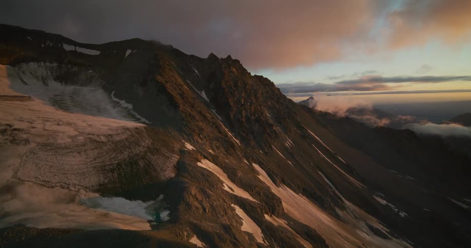 A helicopter flies away over a vast, snow-capped mountain range under a dramatic sunset sky