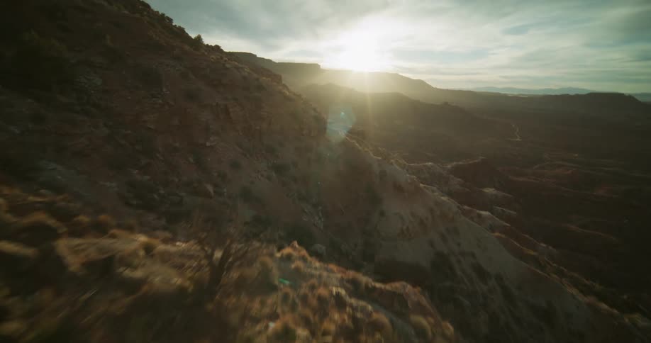 A fast-paced first-person view drone flight soars low over a rugged canyon hillside revealing a vast desert landscape at golden hour