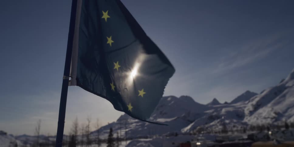 The Alaskan state flag waves in slow motion, silhouetted against the bright sun with snowy mountains in the background
