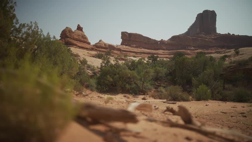 A low-angle, wide shot captures the stark beauty of a dry desert wash leading towards majestic red rock formations under a hazy sky