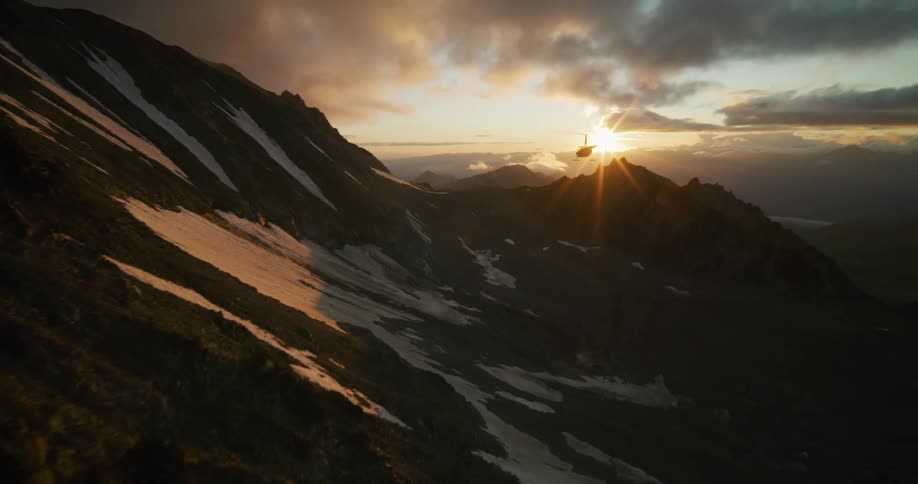 An aerial view follows a helicopter as it flies over a vast, snow-capped mountain range during a stunning golden sunset
