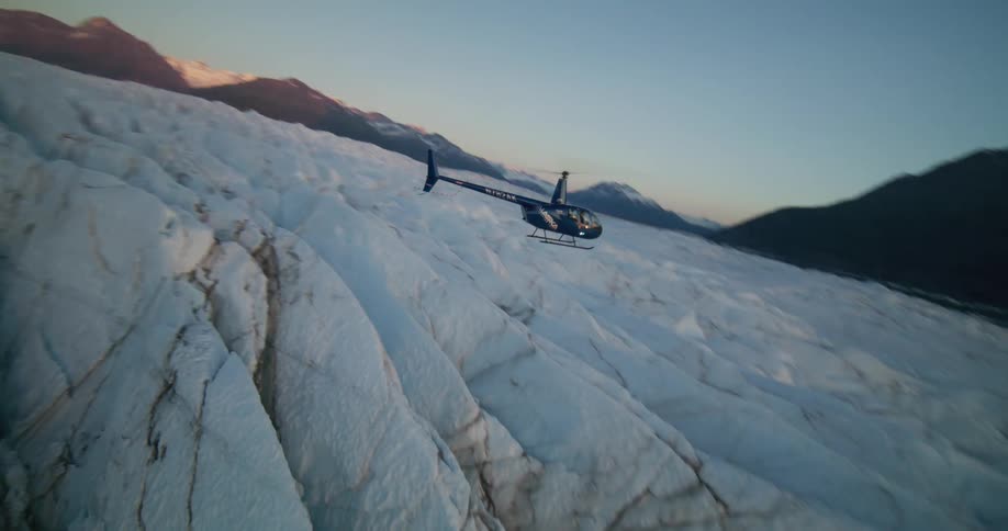 A dramatic vertical aerial shot follows a helicopter flying low over a vast, crevassed glacier at sunrise