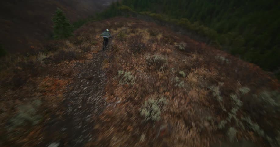An exhilarating aerial tracking shot follows a mountain biker navigating a rugged trail along a vast mountain ridgeline on a cloudy day
