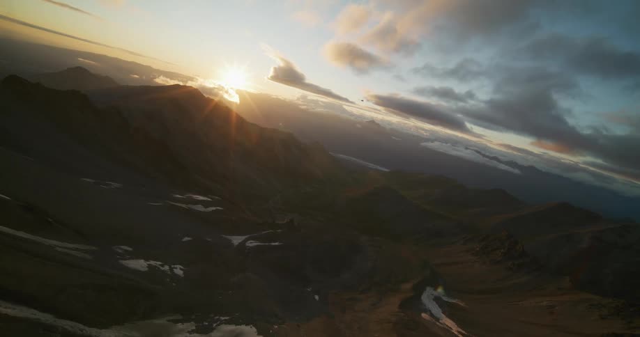 An epic, canted aerial shot reveals a majestic mountain range during a golden sunrise, with a helicopter flying in the distance