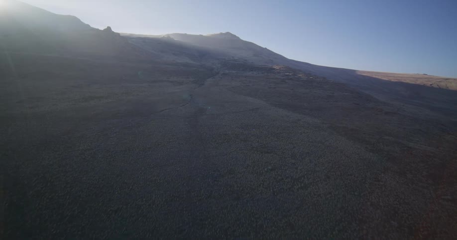Aerial footage glides over a winding road through a desolate, scrub-covered landscape towards a sunlit mountain range