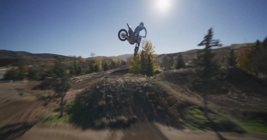 Aerial FPV drone follows a motocross rider as they speed along a dirt track, launching off a jump for a huge whip trick in slow motion