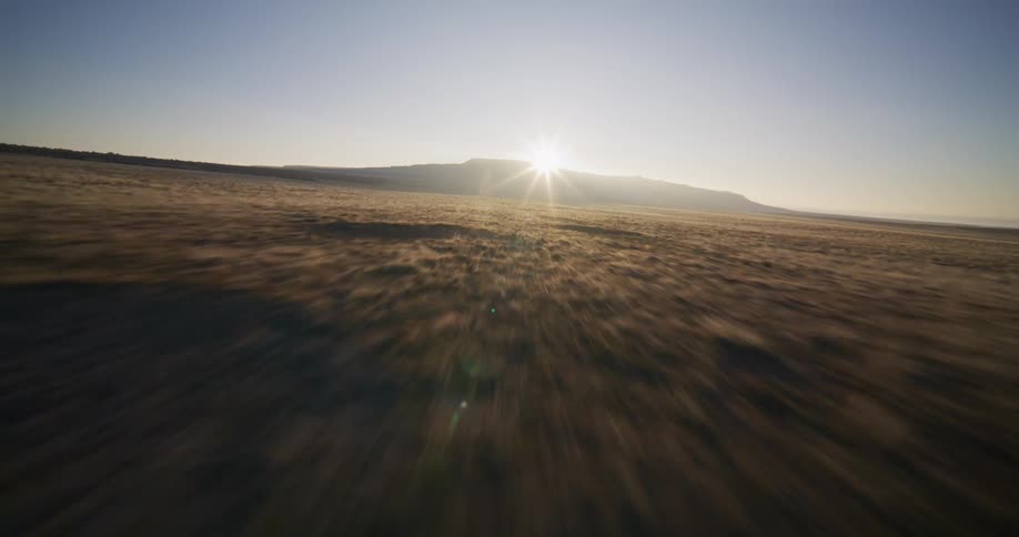 An exhilarating FPV drone flies low and fast across a vast, arid landscape towards a mesa backlit by the rising sun