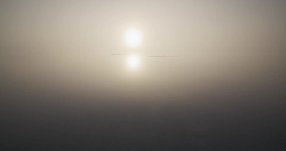 Ethereal low-angle aerial shot gliding over a barren, misty shoreline and calm, reflective water towards a hazy sunrise