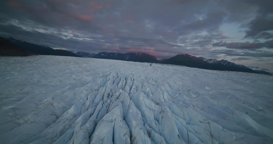 A breathtaking aerial flight over a vast, deeply crevassed glacier landscape under a dramatic sunset sky