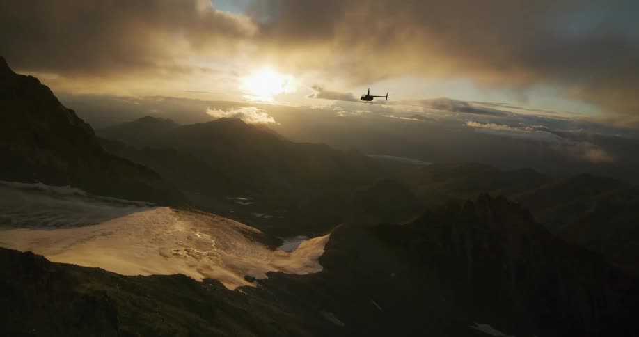 A helicopter flies in slow motion through a majestic mountain range, silhouetted against a dramatic golden sunrise