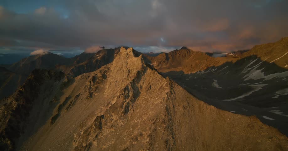 A breathtaking aerial view follows a helicopter flying over a majestic, rocky mountain range during a golden sunset